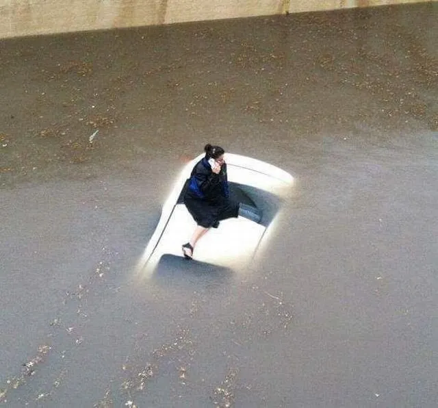 A photograph of a woman stranded on top of a car hood in the middle of a flood. She appears to be on her phone making a call.