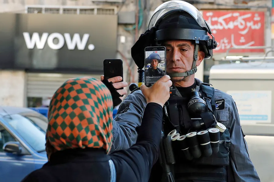 A photograph of a police officer in riot gear and a woman in a head scarf who appear to be standing off. Each person is pointing their smart phone at the other, presumably recording one another.