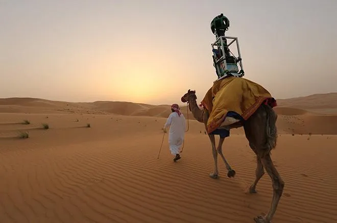 A photograph of a man and a camel in what appears to be the Saharan desert. The camel has an apparatus strapped atop its back, which looks like a Google Maps photo sphere, for recording the surrounding environment.
